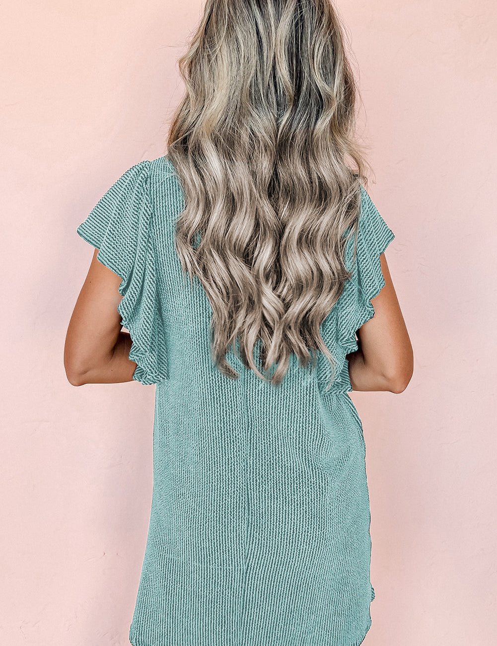 Donna con capelli ondulati biondi indossa un abito verde menta a coste con maniche corte volant, sfondo rosa. Moda estiva, abbigliamento casual.