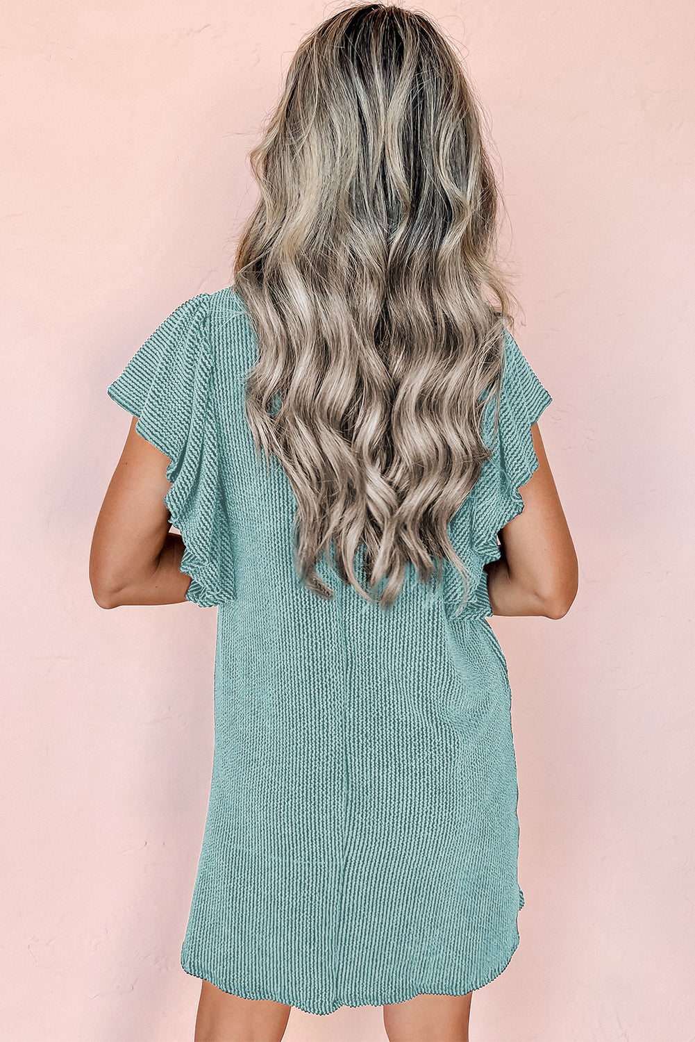 Donna con capelli ondulati biondi indossa un abito verde menta a coste con maniche corte volant, sfondo rosa. Moda estiva, abbigliamento casual.