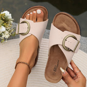 Sandali donna beige con fibbia grande, suola in sughero, indossati su tappeto bianco. Design estivo elegante, perfetti per moda casual e comfort.