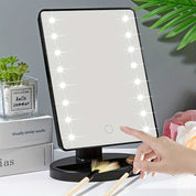 Specchio cosmetico LED rettangolare con base nera, circondato da luci brillanti, piante decorative e pennelli da trucco su tavolo da toeletta.