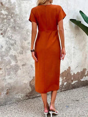 Abito midi arancione donna, maniche corte, schiena scoperta, stile casual estivo, indossato con sandali bianchi, sfondo muro rustico.