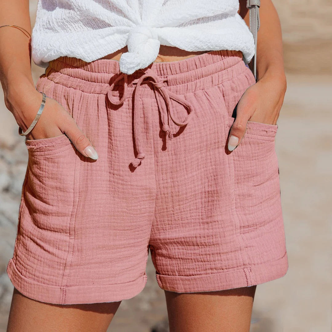 Donna indossa shorts rosa in cotone con tasche, laccio in vita, abbinati a top bianco annodato. Moda estiva casual, abbigliamento donna.