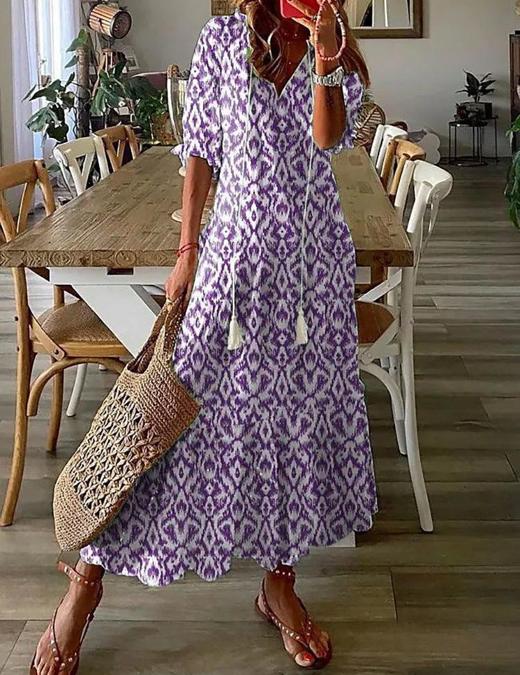 Donna con abito lungo boho viola e bianco, motivo etnico, in piedi in sala da pranzo. Borsa di paglia intrecciata, sandali marroni. Arredamento moderno.