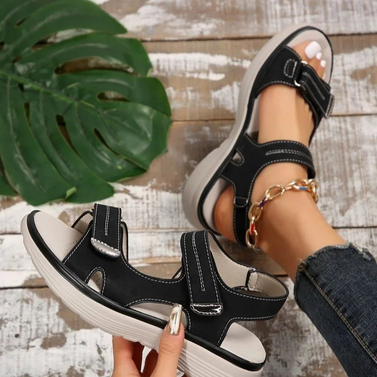 Sandali neri da donna con suola spessa, cinturini regolabili, indossati su pavimento in legno con foglia verde decorativa. Moda estiva casual.
