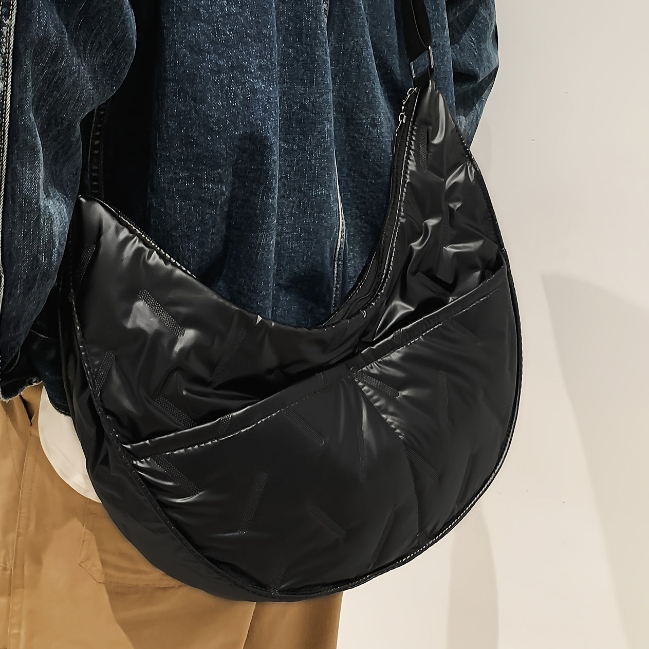 Borsa a tracolla nera in nylon lucido, design moderno e spazioso, indossata su giacca di jeans e pantaloni beige. Accessorio moda unisex.