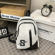 Zaino bianco e nero con logo "S", tasche multiple, su pavimento a scacchi. Orologio nero e scatola industriale sullo sfondo.