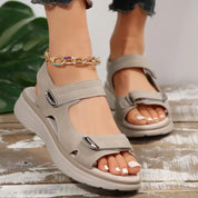Sandali estivi da donna beige con zeppa, chiusura a strappo, suola antiscivolo, indossati su pavimento in legno, pianta verde sullo sfondo.
