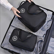 Utente che organizza valigia con borse da viaggio nere, logo "M", su camicie a righe. Accessori viaggio, organizzazione bagagli, valigia ordinata.