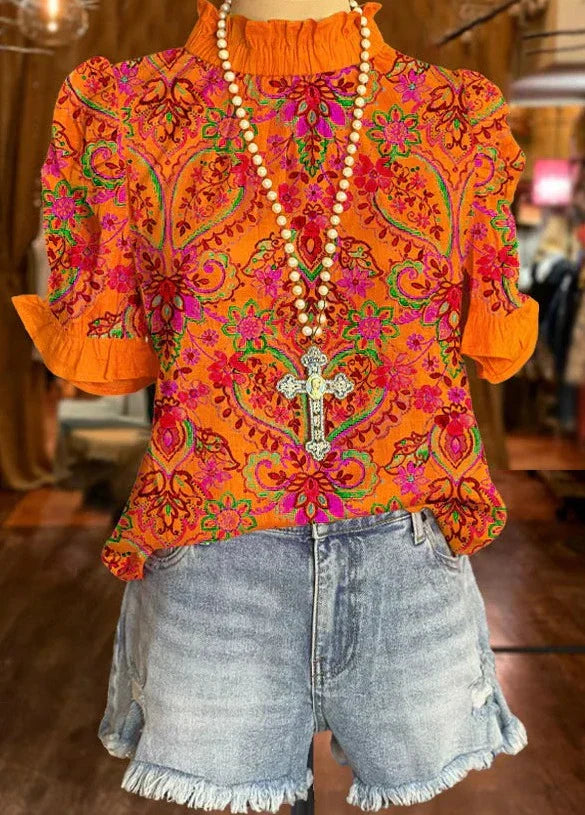 Blusa arancione con motivi floreali rosa e verdi, collana con croce, abbinata a shorts di jeans sfrangiati. Moda estiva, abbigliamento casual donna.