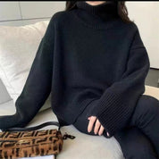 Maglione nero a collo alto in lana, stile oversize, abbinato a borsa Fendi marrone con motivo iconico, su sfondo di divano moderno.