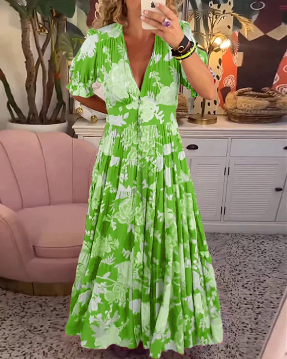Abito lungo verde con stampa floreale bianca, scollo a V, maniche corte, indossato da una donna in un salotto moderno con poltrona rosa e piante.