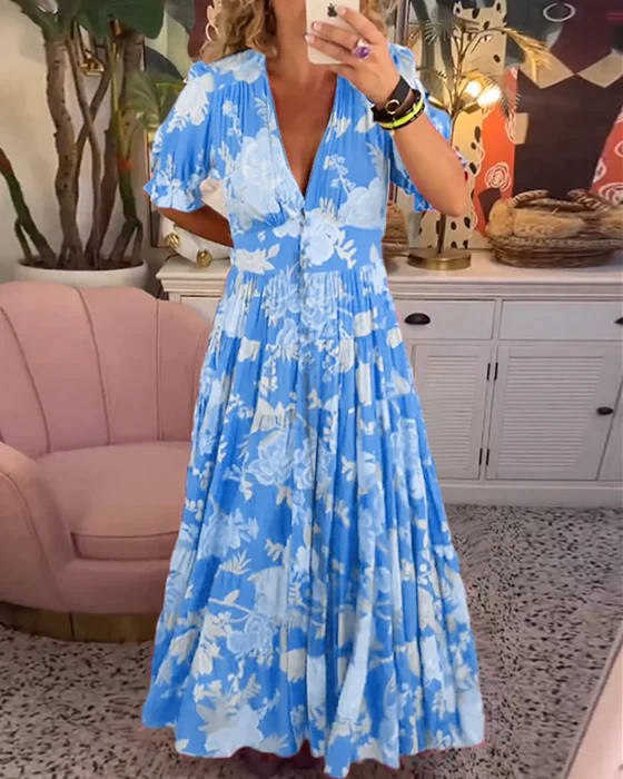 Abito lungo estivo blu con stampa floreale bianca, scollo a V, maniche corte, indossato da una donna in un salotto moderno con poltrona rosa.