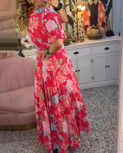 Abito lungo floreale rosso e rosa, donna di spalle in salotto elegante con mobili bianchi e decorazioni artistiche. Moda estiva, stile boho chic.