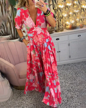 Abito lungo floreale rosso con scollo a V, maniche corte e tasche, indossato da una donna in un salotto elegante con divano rosa e decorazioni tropicali.