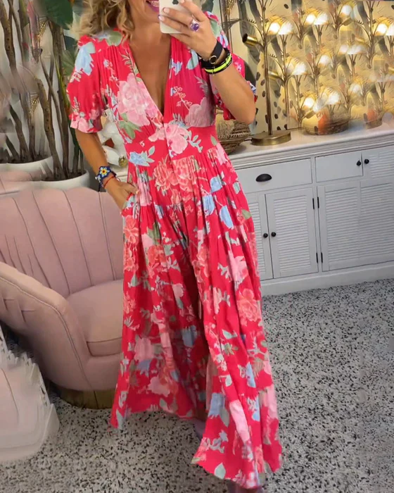 Abito lungo floreale rosso con scollo a V, maniche corte e tasche, indossato da una donna in un salotto elegante con divano rosa e decorazioni tropicali.