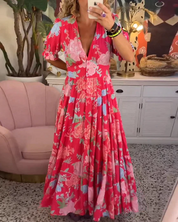 Abito lungo floreale rosa con scollo a V, maniche corte, indossato da una donna in un salotto elegante con poltrona rosa e mobili bianchi.