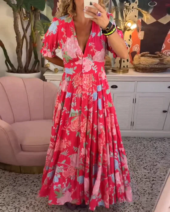 Abito lungo floreale rosa con scollo a V, maniche corte, indossato da una donna in un salotto elegante con poltrona rosa e mobili bianchi.
