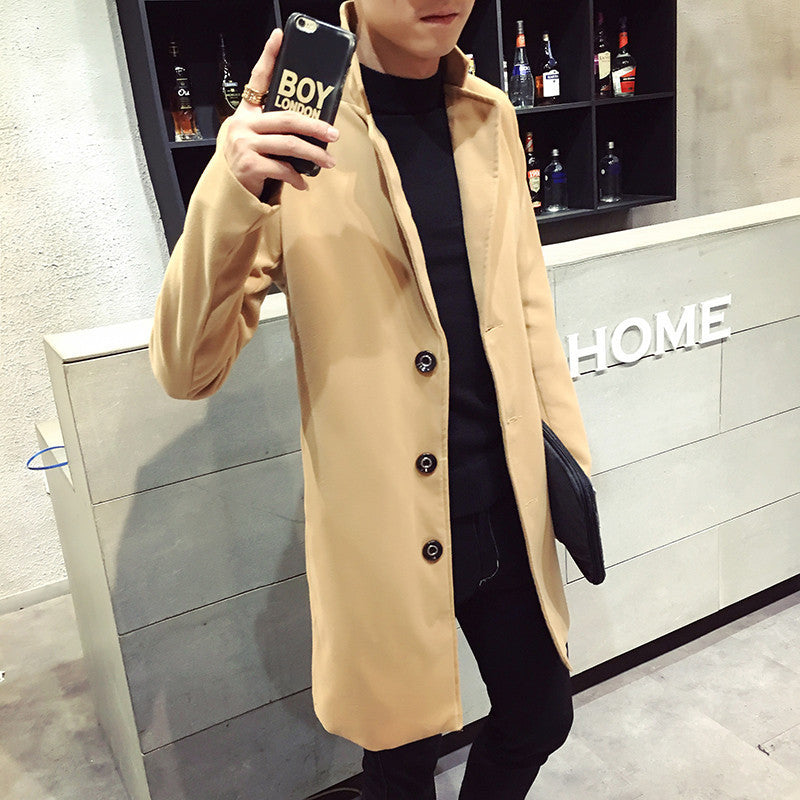 Uomo con cappotto beige elegante, smartphone in mano, sfondo con bottiglie su scaffali. Moda maschile, abbigliamento autunnale, stile urbano.