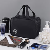 Borsa da toilette nera con logo "M", doppia cerniera, circondata da accessori da bagno: spazzolino, crema, rasoio, tazza termica, sapone.