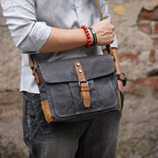 Uomo con borsa a tracolla in tela grigia, dettagli in pelle marrone, indossa camicia azzurra e bracciale rosso. Stile casual urbano.