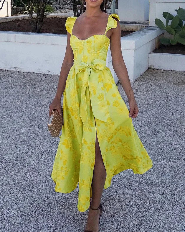 Donna con abito giallo floreale estivo, spacco frontale, cintura in vita, sandali marroni, borsa intrecciata. Look elegante per eventi estivi.