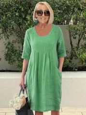 Donna con abito verde in lino, occhiali da sole, orecchini turchesi, borsa nera e fiori bianchi. Moda estiva casual, look elegante e fresco.