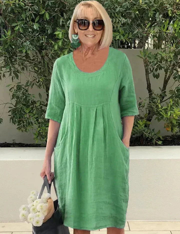 Donna con abito verde in lino, occhiali da sole, orecchini turchesi, borsa nera e fiori bianchi. Moda estiva casual, look elegante e fresco.