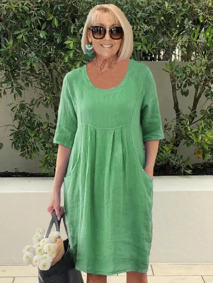 Donna con abito verde in lino, occhiali da sole, orecchini turchesi, borsa nera e fiori bianchi. Moda estiva casual, look elegante e fresco.