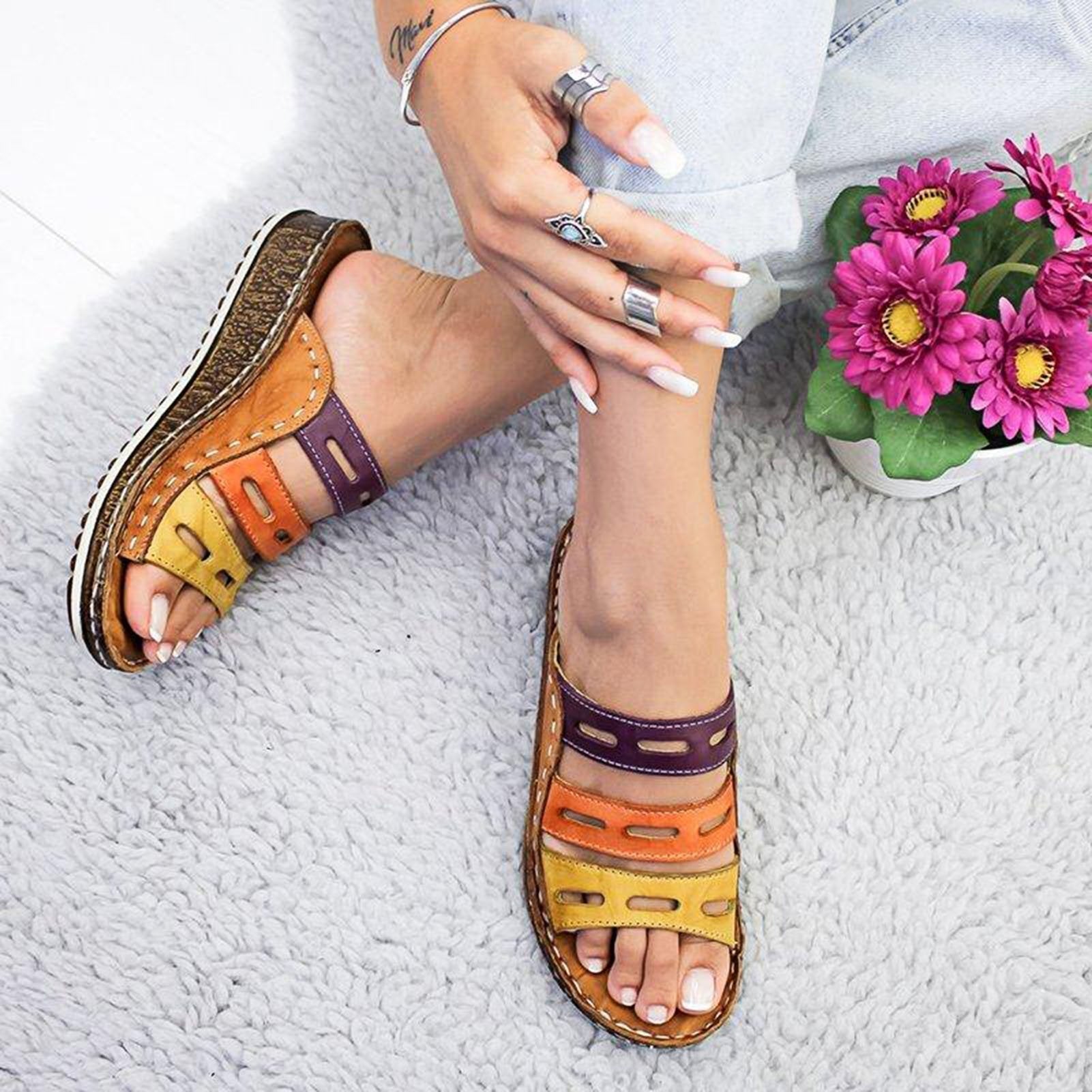 Donna con sandali multicolore in pelle su tappeto grigio, fiori rosa accanto. Scarpe estive moda, comfort, stile casual.