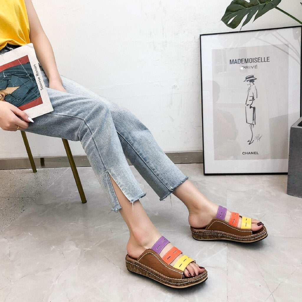 Donna seduta con jeans chiari e sandali colorati su suola in corda, accanto a un quadro appoggiato a parete. Moda estiva casual, calzature trendy.