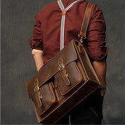 Borsa a tracolla in pelle marrone, design vintage, con doppie tasche frontali e fibbie, indossata da uomo con camicia bordeaux e bretelle.