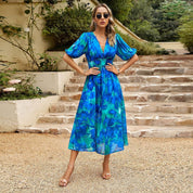 Donna con abito lungo floreale blu-verde, occhiali da sole, sandali beige, su scala esterna. Moda estiva elegante, abbigliamento femminile trendy.