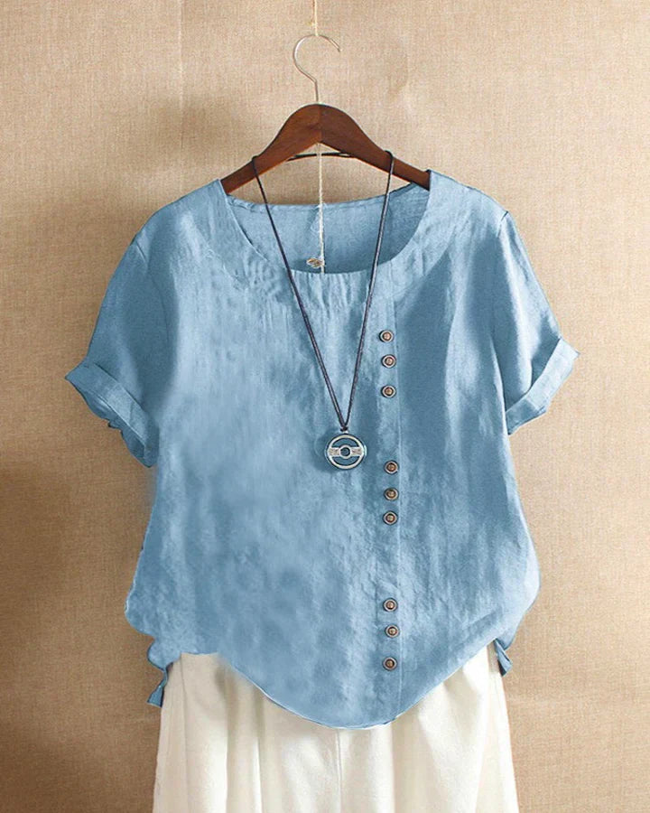 Blusa donna in lino azzurro con bottoni asimmetrici, maniche corte e scollo rotondo, appesa su gruccia. Stile casual estivo.