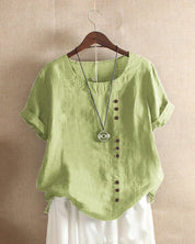 Blusa verde in lino con bottoni asimmetrici, maniche corte e collana pendente, appesa su gruccia. Moda estiva casual donna.