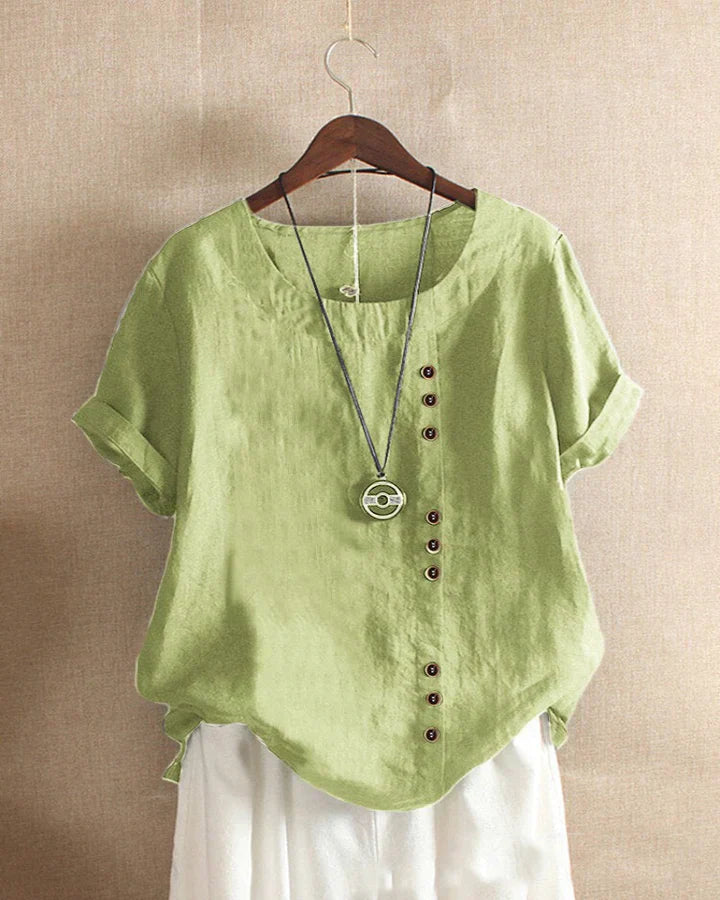 Blusa verde in lino con bottoni asimmetrici, maniche corte e collana pendente, appesa su gruccia. Moda estiva casual donna.