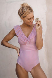 Donna con costume intero a righe rosa e bianche, scollo a V e volant, tiene un fiore. Moda mare estiva, abbigliamento da spiaggia elegante.