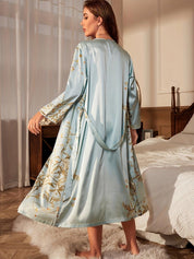 Donna in camera da letto indossa vestaglia di seta azzurra con motivi floreali dorati, elegante e lussuosa, su tappeto bianco soffice.