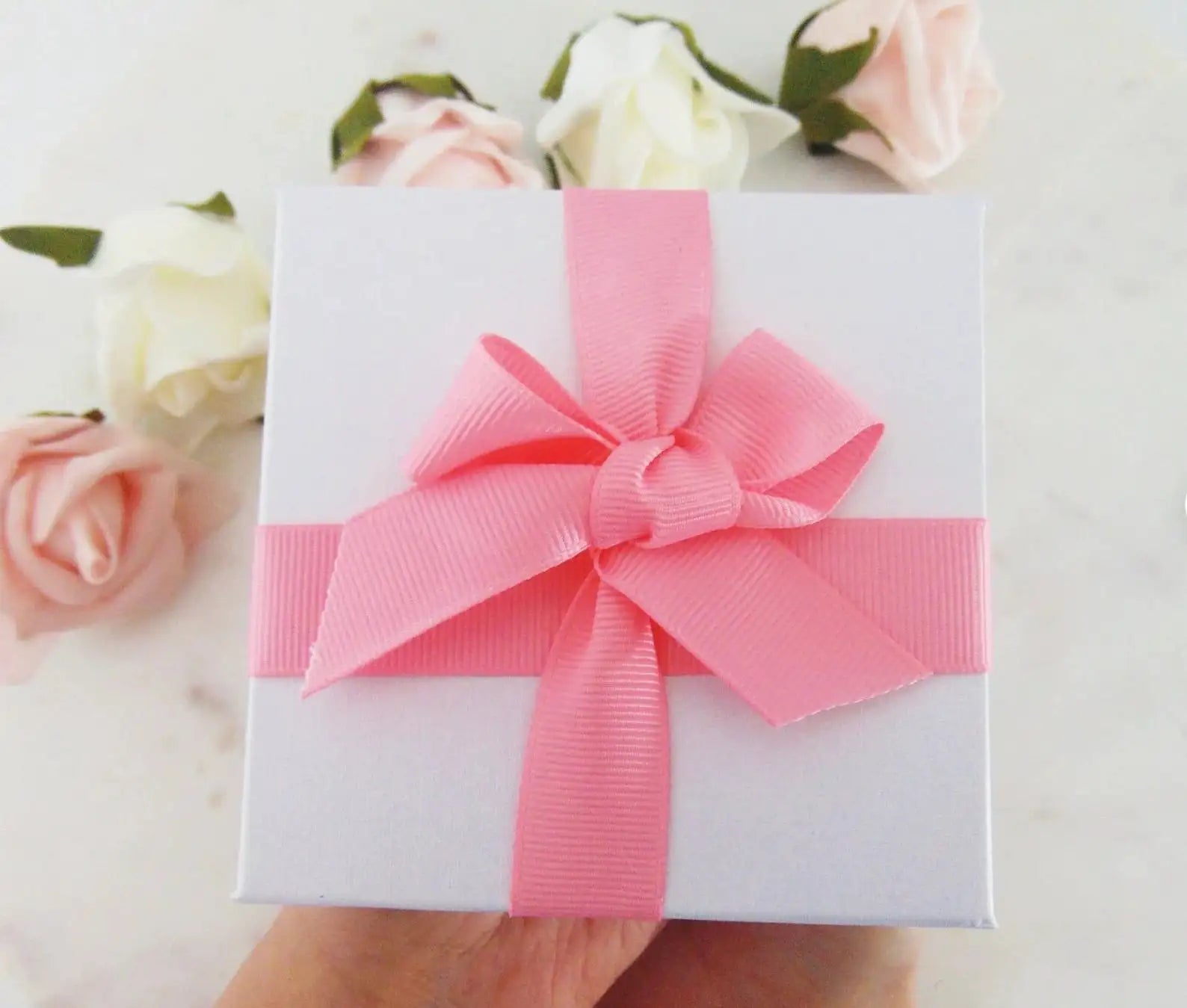 Confezione regalo bianca con fiocco rosa, circondata da rose bianche e rosa. Idea regalo elegante, perfetta per occasioni speciali e matrimoni.