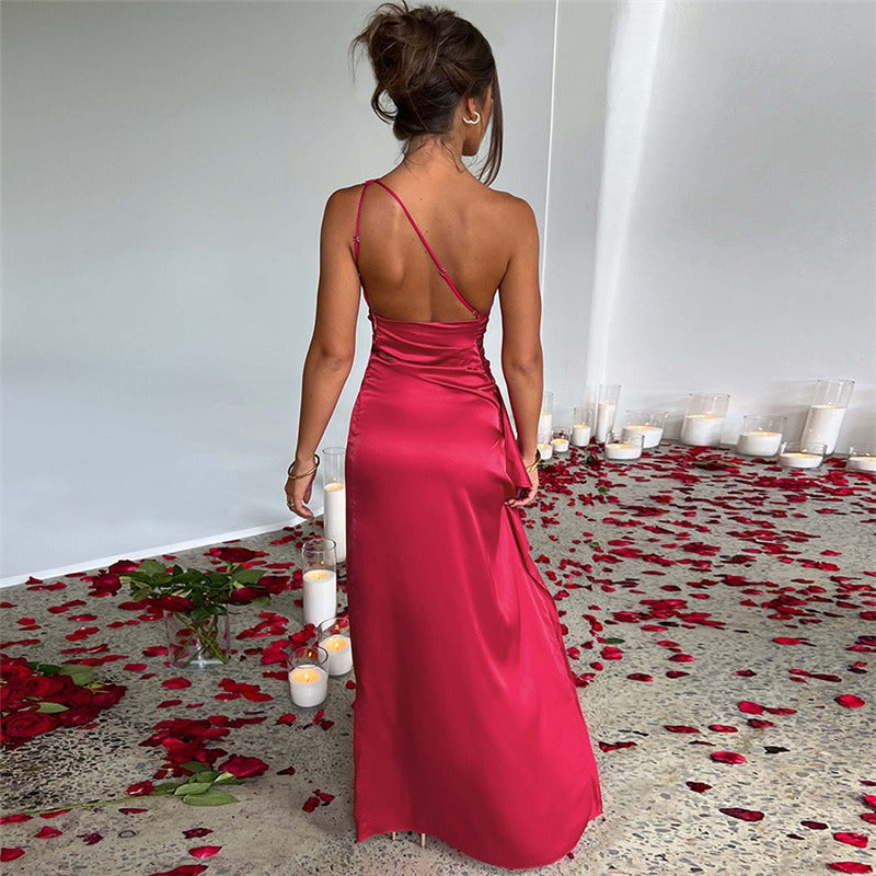 Abito lungo rosso elegante in raso, spallina sottile, schiena scoperta, circondato da petali di rosa e candele bianche su pavimento grigio.