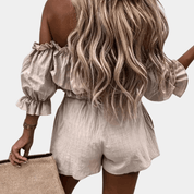 Donna con capelli biondi lunghi indossa tuta corta beige con spalle scoperte e volant, tiene borsa in paglia. Moda estiva elegante e casual.