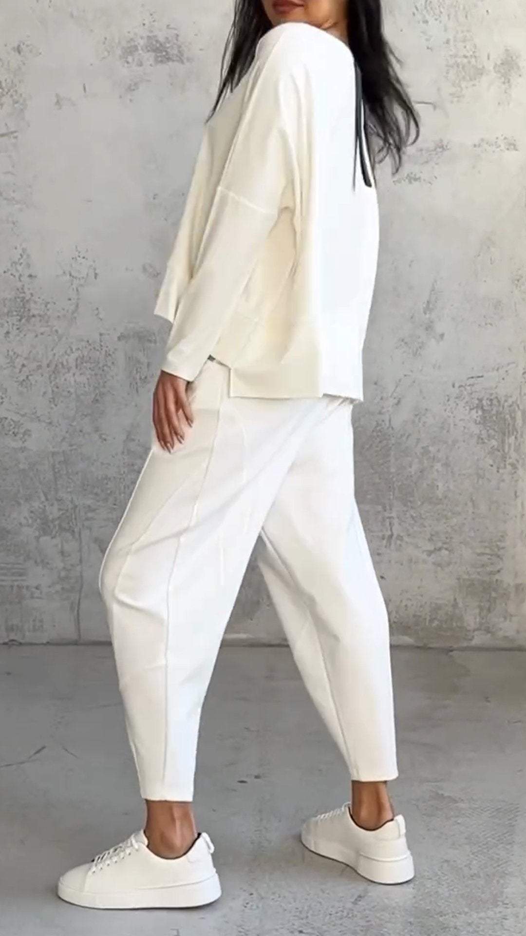 Donna in abbigliamento casual bianco, indossa maglia e pantaloni larghi, scarpe da ginnastica bianche, posa su sfondo grigio. Moda minimalista elegante.