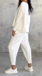 Donna in abbigliamento casual bianco, indossa maglia e pantaloni larghi, scarpe da ginnastica bianche, posa su sfondo grigio. Moda minimalista elegante.