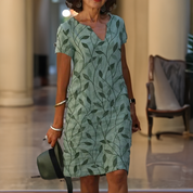 Abito estivo verde con motivo a foglie, maniche corte, indossato da donna in interno elegante. Accessori: cappello verde, bracciali. Moda femminile.