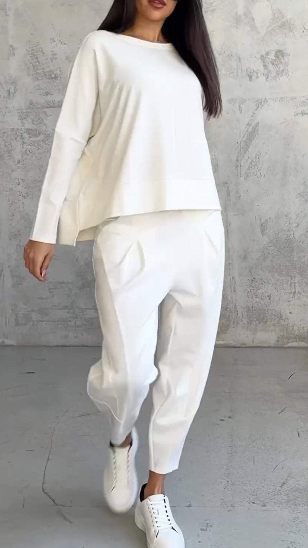 Donna con abbigliamento casual bianco: maglia a maniche lunghe, pantaloni larghi, sneakers. Moda minimalista, stile moderno, sfondo grigio.