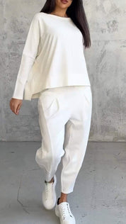 Donna con abbigliamento casual bianco: maglia a maniche lunghe, pantaloni larghi, sneakers. Moda minimalista, stile moderno, sfondo grigio.
