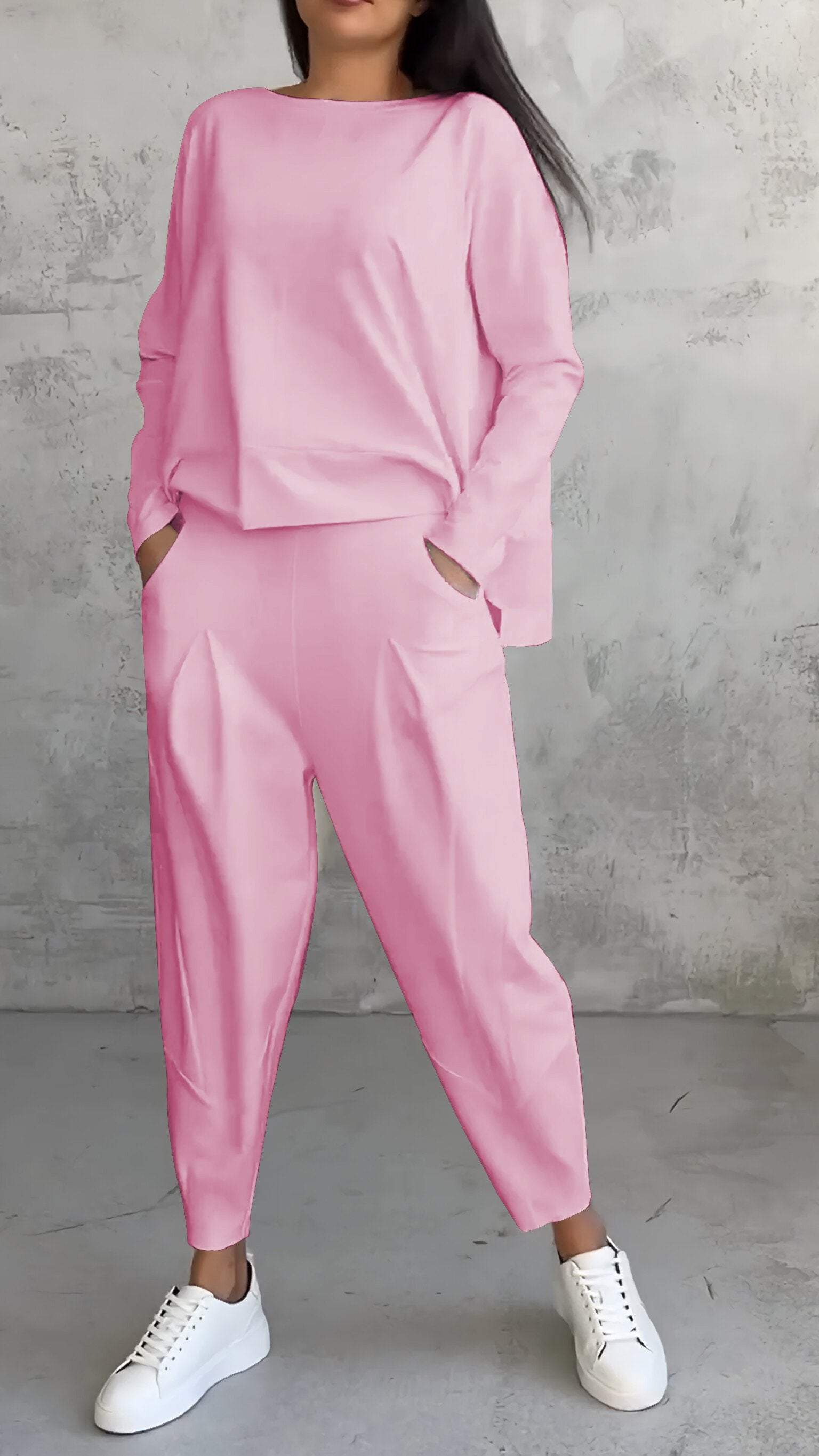 Completo donna rosa pastello, maglia e pantaloni larghi, tasche laterali, abbinato a sneakers bianche, sfondo grigio minimalista.