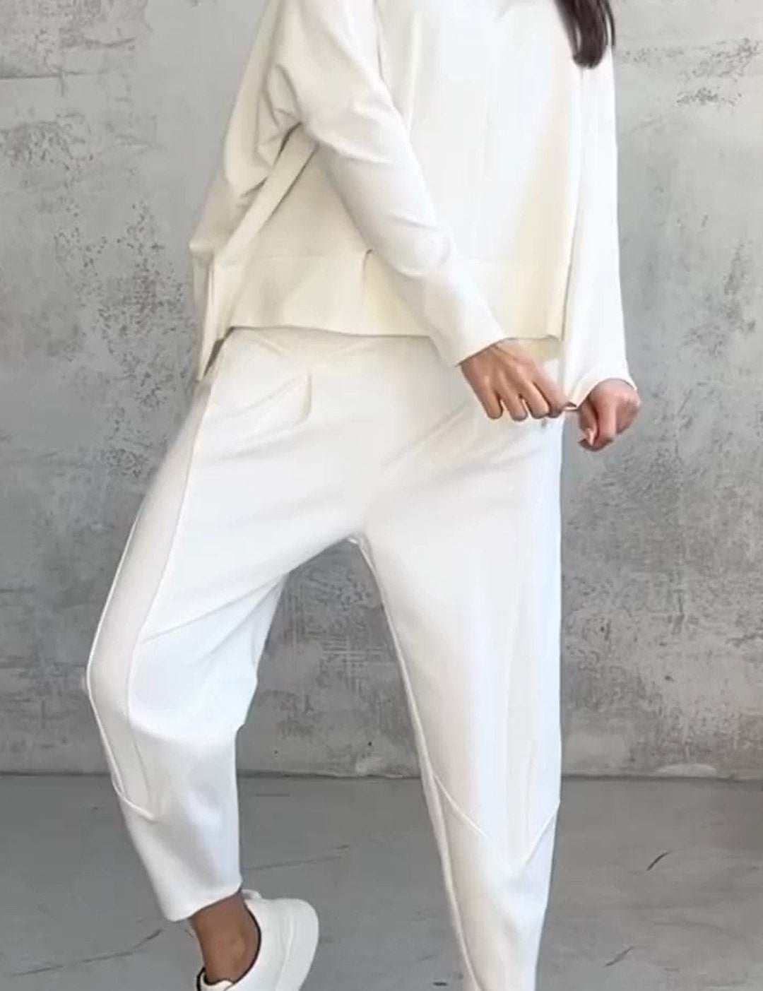 Donna in completo bianco casual, composto da maglia e pantaloni larghi, con sneakers bianche. Look minimalista e moderno su sfondo grigio.