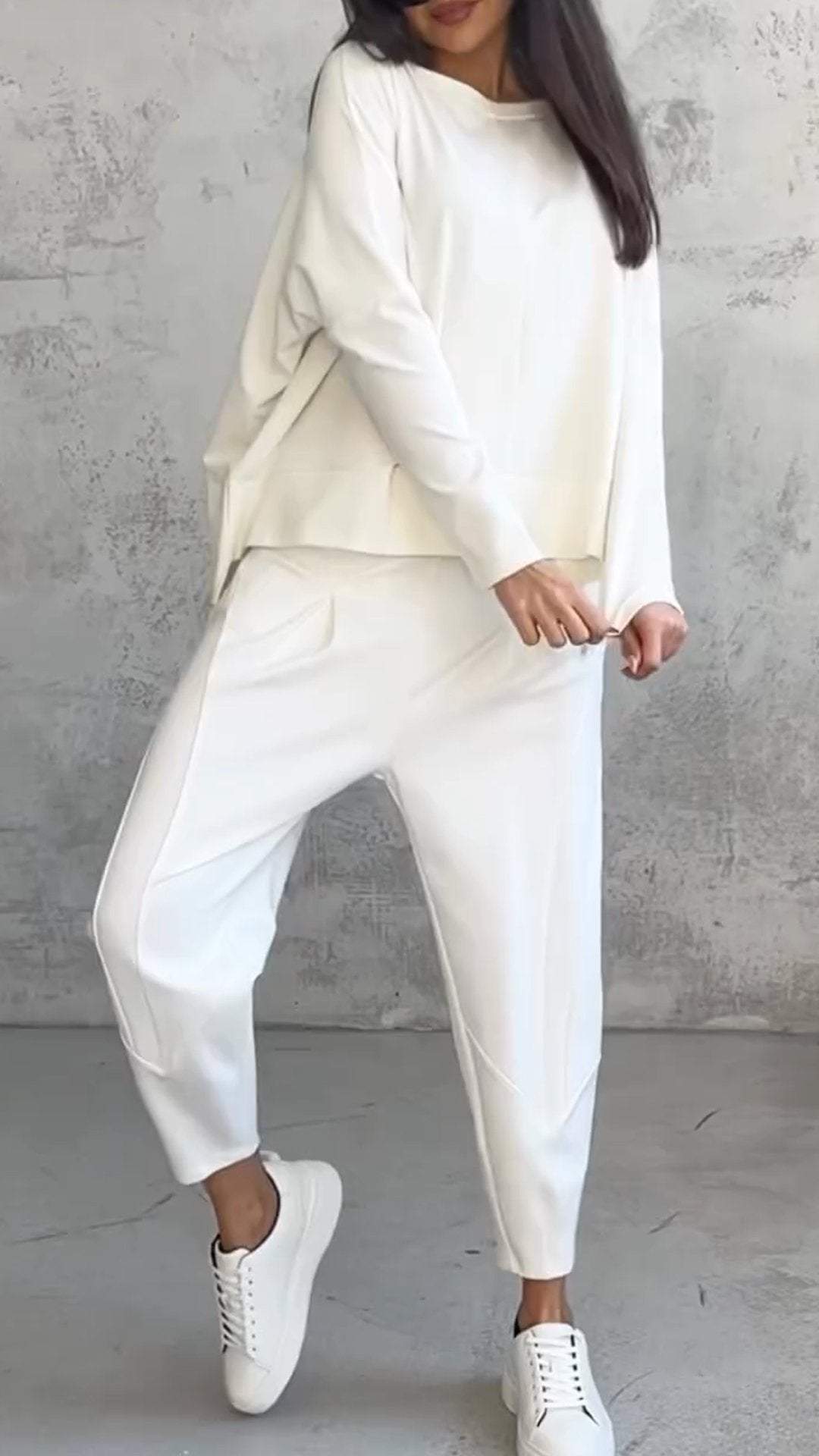 Donna in completo bianco casual, composto da maglia e pantaloni larghi, con sneakers bianche. Look minimalista e moderno su sfondo grigio.
