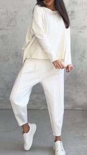 Donna in completo bianco casual, composto da maglia e pantaloni larghi, con sneakers bianche. Look minimalista e moderno su sfondo grigio.