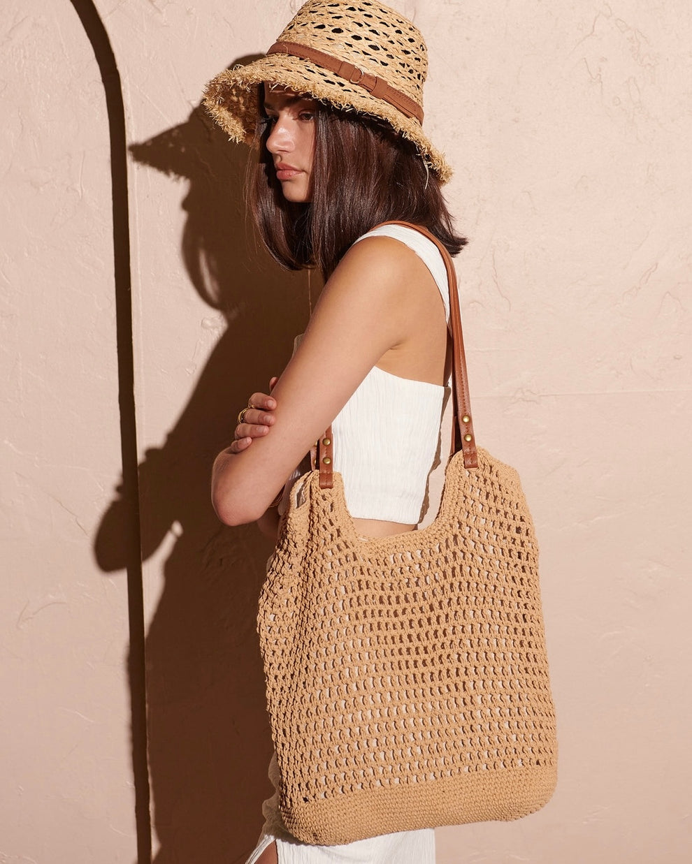 Donna con cappello di paglia e borsa intrecciata beige, abito bianco estivo, sfondo rosa. Moda estiva, accessori eleganti, stile boho chic.